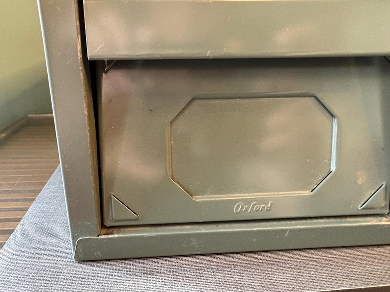 May include: Close-up of a vintage olive green metal file cabinet drawer. The drawer has a recessed panel with the name "Oxford" embossed on it. The cabinet appears to be made of sturdy metal.