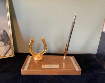 Könnte beinhalten: Ein Schreibtisch-Organizer aus Holz mit einem goldenen Hufeisen und einem silbernen Stifthalter. Der Schreibtisch-Organizer hat eine Plakette mit der Aufschrift "Viel Glück immer".