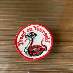 Puede incluir: Parche redondo con fondo blanco y borde rojo. Presenta una serpiente negra y roja con las palabras "Tread on Yourself" en letras rojas. La serpiente está enrollada y tiene la lengua afuera.