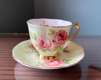 May include: A delicate teacup and saucer set with a floral design. The cup and saucer are both light green with a gold trim. The floral design features pink and red roses with green leaves.