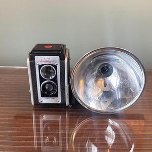 Peut inclure: Un appareil photo reflex bi-objectif vintage Kodak Duaflex II avec un corps noir texturé et un grand réflecteur de flash circulaire en chrome poli fixé sur le côté. La face avant présente deux objectifs en verre et le texte Duaflex II Kodak.