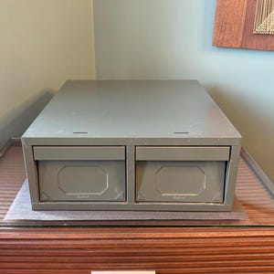 May include: A two-drawer metal storage cabinet in a muted green colour. The cabinet sits on a wooden surface with a glass top. The drawers have a rectangular design with a central, octagonal indentation. The cabinet is a storage solution.