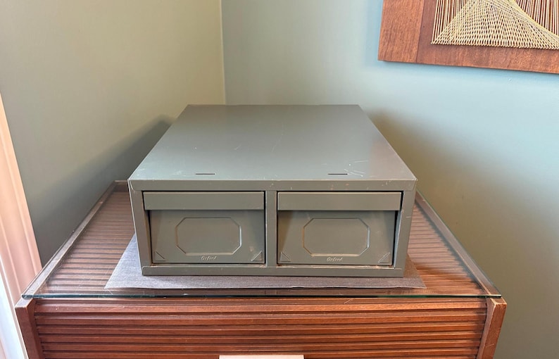 May include: A two-drawer metal storage cabinet in a muted green colour. The cabinet sits on a wooden surface with a glass top. The drawers have a rectangular design with a central, octagonal indentation. The cabinet is a storage solution.