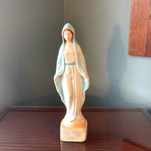 Könnte beinhalten: Eine Vintage-Statue der Jungfrau Maria, mit einem hellblauen und weißen Gewand und passender Kopfbedeckung. Die Statue hat ein pfirsichfarbenes Gesicht und Hände und steht auf einem strukturierten, beigefarbenen Sockel. Die religiöse Figur wird auf einer dunkelbraunen Oberfläche ausgestellt.