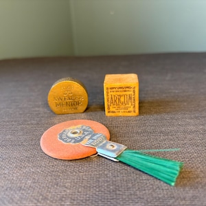 May include: Three vintage items are displayed on a textured surface. A small, round, gold-colored object with text, a small orange box with text, and a round, orange object with a green brush attached.