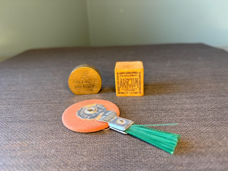 May include: Three vintage items are displayed on a textured surface. A small, round, gold-colored object with text, a small orange box with text, and a round, orange object with a green brush attached.