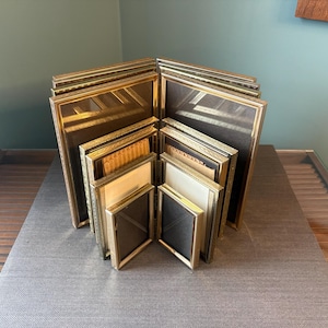 May include: A collection of vintage gold-framed picture frames of various sizes, arranged in a fan shape. The frames have a classic design with ornate detailing and are displayed on a grey surface.