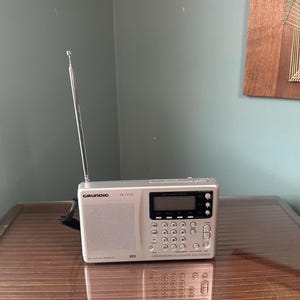 以下が含まれることがあります： デジタルディスプレイ、テンキー、長く伸びる金属製アンテナを備えたシルバーのGrundig YB 400PEポータブルラジオ。フロントパネルの左側にはスピーカーグリルがあり、チューニングや音量調整用のさまざまなコントロールボタンが配置されています。