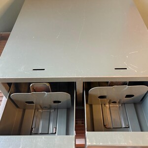 May include: A grey metal cabinet with two open drawers. Each drawer has a metal divider and a small, rectangular compartment. The cabinet has a slightly distressed appearance.