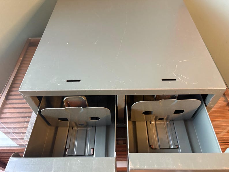May include: A grey metal cabinet with two open drawers. Each drawer has a metal divider and a small, rectangular compartment. The cabinet has a slightly distressed appearance.