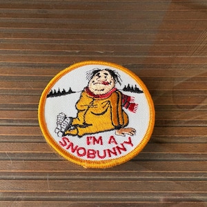 May include: Round embroidered patch featuring a cartoon figure in a yellow outfit and red scarf, with the text "I'M A SNOBUNNY". The patch has a white background, an orange border, and a small black mountain range.