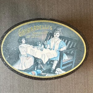 May include: Vintage oval tin featuring a charming illustration of two young girls at a tea party, with a teddy bear and a dog. The tin is labeled "Ghirardelli's Chocolate de Cocoa" in gold lettering.