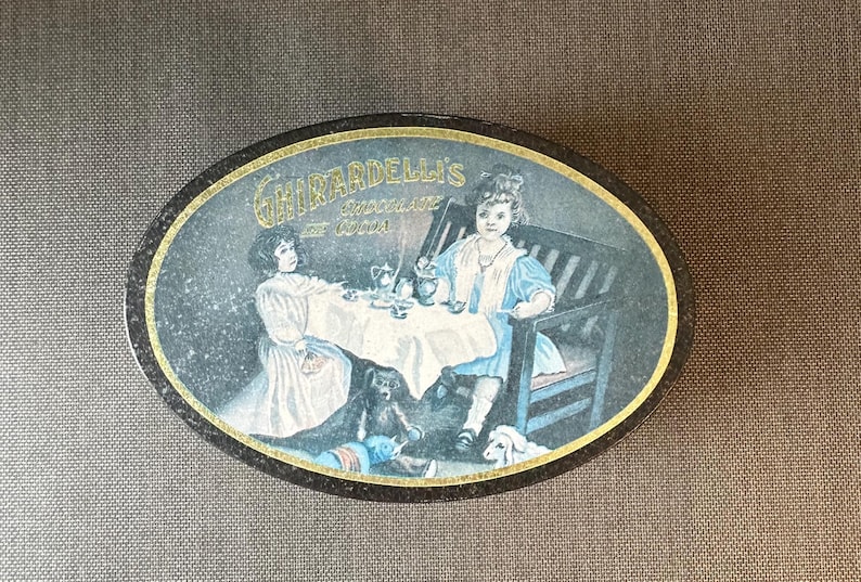 May include: Vintage oval tin featuring a charming illustration of two young girls at a tea party, with a teddy bear and a dog. The tin is labeled "Ghirardelli's Chocolate de Cocoa" in gold lettering.