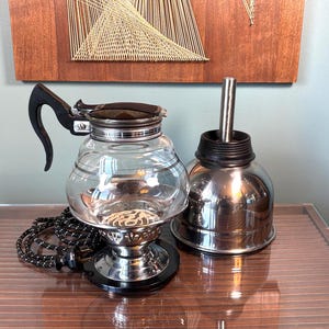 May include: A vintage electric glass coffee percolator with a black handle and base, featuring a clear glass carafe, a metallic heating element, and a separate stainless steel lid and internal stem assembly, accompanied by a black and white power cord.