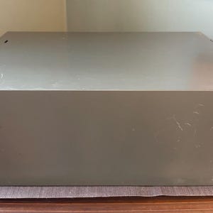 May include: A large, rectangular, olive-green metal box with a slightly textured surface. The box has a simple, utilitarian design with a few small holes on the top. It sits on a brown and beige surface.