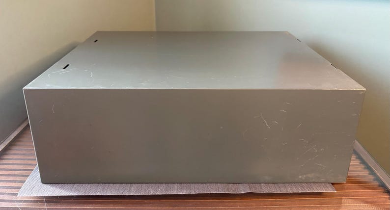 May include: A large, rectangular, olive-green metal box with a slightly textured surface. The box has a simple, utilitarian design with a few small holes on the top. It sits on a brown and beige surface.