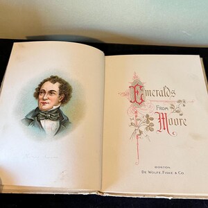 May include: An open book with a portrait of a man on the left page and the title "Emeralds from Moore" on the right page. The book is bound in cream-colored paper with gold accents.