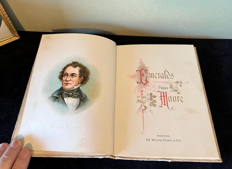 May include: An open book with a portrait of a man on the left page and the title "Emeralds from Moore" on the right page. The book is bound in cream-colored paper with gold accents.