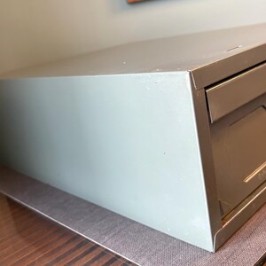 May include: A light grey metal storage cabinet with two drawers. The cabinet has a rectangular shape and is sitting on a brown surface. The drawers have a geometric design.