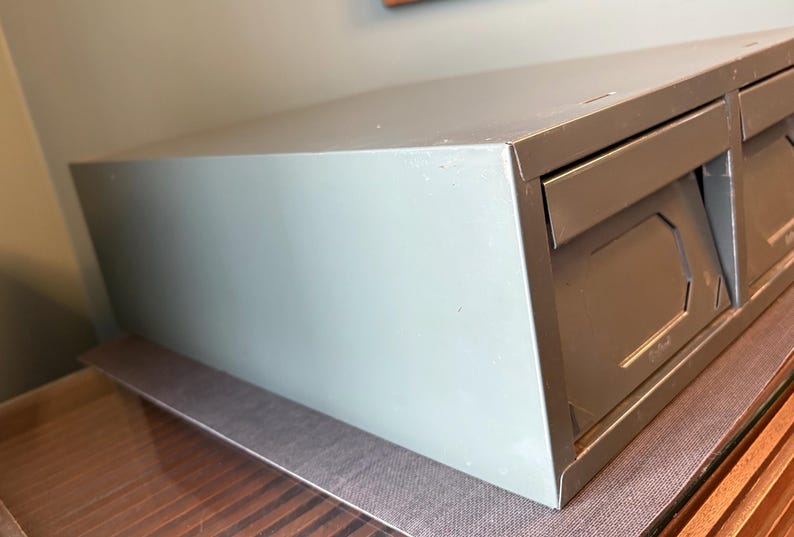 May include: A light grey metal storage cabinet with two drawers. The cabinet has a rectangular shape and is sitting on a brown surface. The drawers have a geometric design.