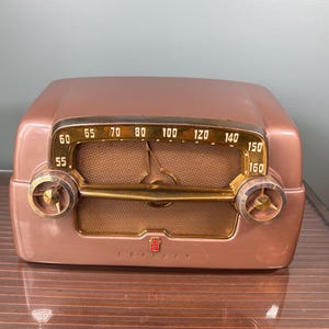 Peut inclure: Une radio de table en or rose métallique dotée d'un cadran doré avec des fréquences allant de 55 à 160. La radio présente deux boutons circulaires sur les côtés, une grille de haut-parleur centrale en tissu marron et un petit logo rouge.