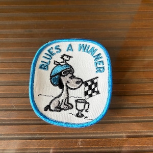 May include: Embroidered patch with a cartoon dog wearing a blue hat and sunglasses, holding a checkered flag. The patch has the text "Blues a Winner" in a blue border, with a trophy at the bottom.