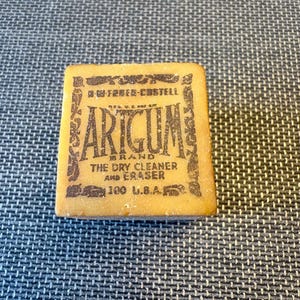 May include: A rectangular Artgum brand dry cleaner and eraser. The eraser is a light tan color with dark brown text that reads "Artgum" and "The Dry Cleaner and Eraser". The text "100 U.S.A." is also visible.