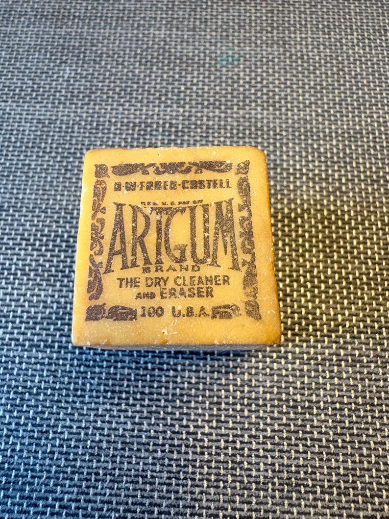 May include: A rectangular Artgum brand dry cleaner and eraser. The eraser is a light tan color with dark brown text that reads "Artgum" and "The Dry Cleaner and Eraser". The text "100 U.S.A." is also visible.