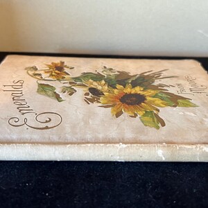 May include: A vintage book titled "Emeralds" with a floral design on the cover. The cover features a bouquet of yellow sunflowers and white flowers with green leaves. The book is bound in white with a cream-colored paper cover.