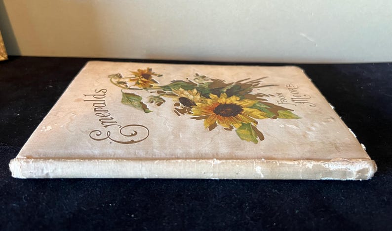 May include: A vintage book titled "Emeralds" with a floral design on the cover. The cover features a bouquet of yellow sunflowers and white flowers with green leaves. The book is bound in white with a cream-colored paper cover.