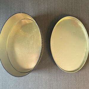 May include: An open, empty, oval-shaped tin with a separate lid. The interior and lid are gold-colored, while the exterior rim of the tin is black. The tin sits on a gray surface.