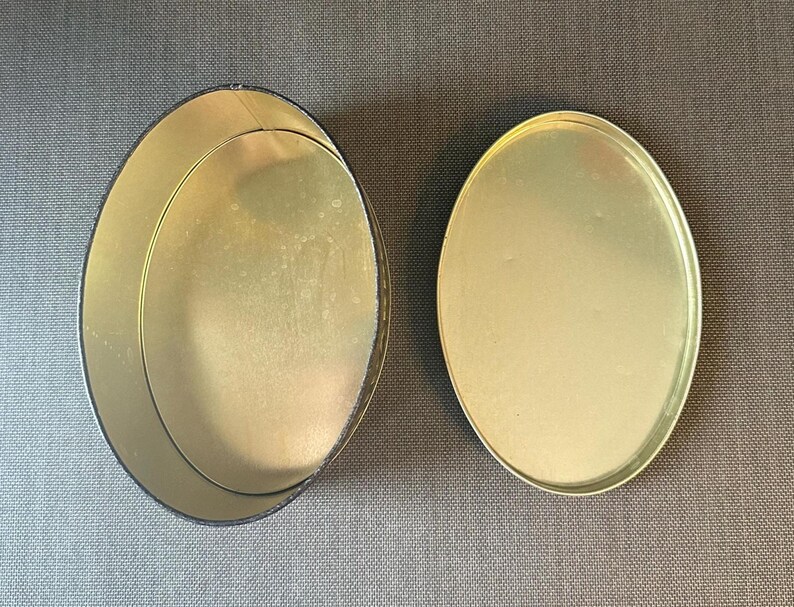 May include: An open, empty, oval-shaped tin with a separate lid. The interior and lid are gold-colored, while the exterior rim of the tin is black. The tin sits on a gray surface.