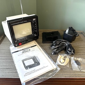 May include: A white and black MTC portable television with a retractable antenna, accompanied by an instruction manual, a black power adapter, a car cigarette lighter power cord, a pair of white wired earbuds, and a small bag of hardware components.