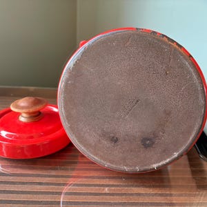 Pu&ograve; includere: Una teiera in smalto rosso con coperchio abbinato e manico in legno. Il coperchio &egrave; rimosso, rivelando l'interno della teiera. La teiera &egrave; appoggiata su una superficie di legno.