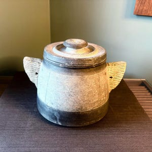 Può includere: Un vaso artigianale, simile alla pietra, con coperchio e due manici strutturati. Il vaso ha una base e un bordo scuri, con una fascia più chiara al centro. Il coperchio ha un piccolo pomello rotondo. Questo pezzo decorativo è adatto per la cucina o l'arredamento della casa.
