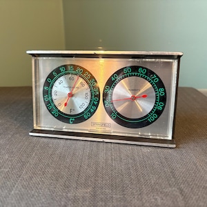 Puede incluir: Un medidor de temperatura y humedad Springfield vintage. El dispositivo plateado tiene dos esferas circulares con marcas negras y verdes. La esfera izquierda muestra la temperatura en Celsius y Fahrenheit, y la derecha la humedad.