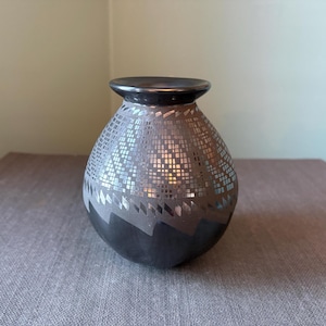 May include: A decorative ceramic vase with a dark gray rim and base. The body of the vase is covered in a mosaic pattern of silver and gray tiles. The vase has a rounded shape and sits on a gray surface.