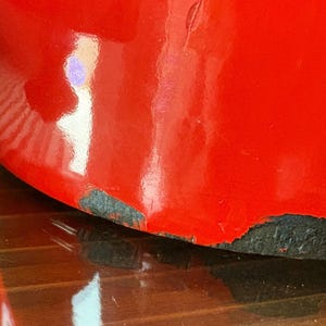 Pu&ograve; includere: Primo piano di un oggetto rosso lucido con segni di usura visibili alla base, che rivelano uno strato inferiore scuro. La superficie riflette la luce, evidenziando la sua forma curva. La forma dell'oggetto &egrave; parzialmente visibile.