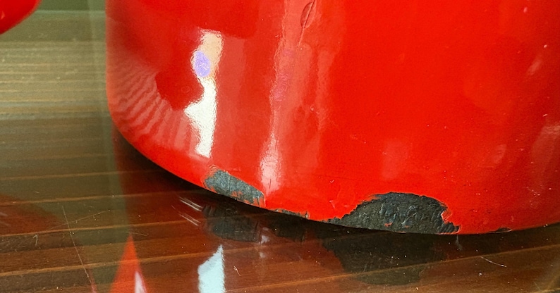 Pu&ograve; includere: Primo piano di un oggetto rosso lucido con segni di usura visibili alla base, che rivelano uno strato inferiore scuro. La superficie riflette la luce, evidenziando la sua forma curva. La forma dell'oggetto &egrave; parzialmente visibile.