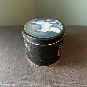 May include: A round black tin with gold lettering and trim. The lid features a detailed illustration. The tin's side displays the word "Gillette" in gold script. The tin is sitting on a textured surface.