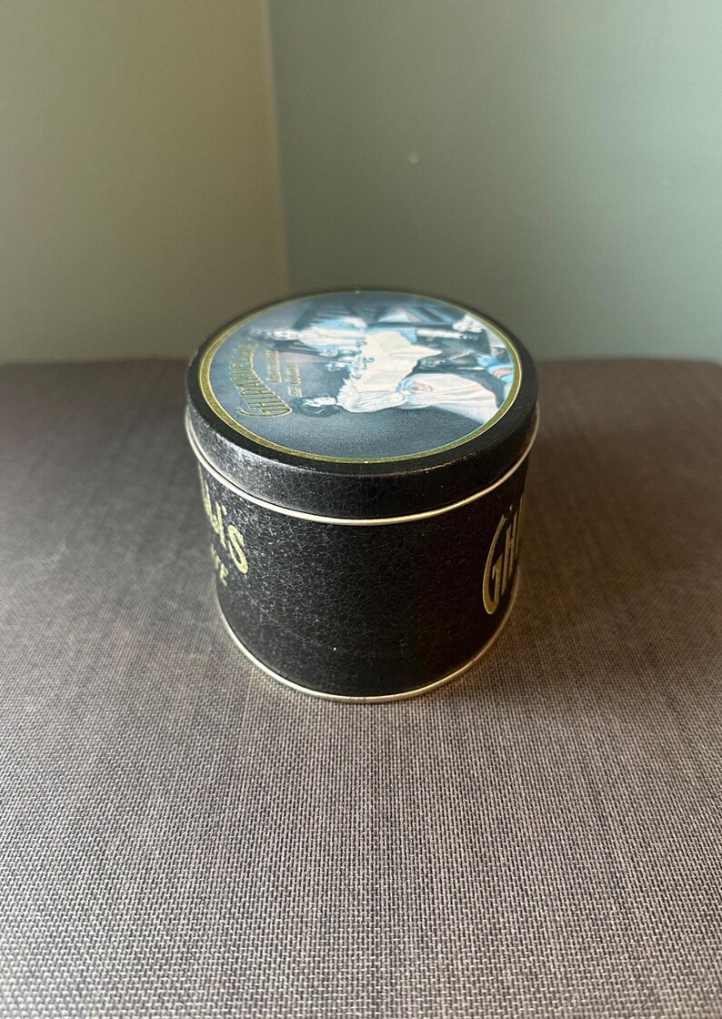 May include: A round black tin with gold lettering and trim. The lid features a detailed illustration. The tin's side displays the word "Gillette" in gold script. The tin is sitting on a textured surface.