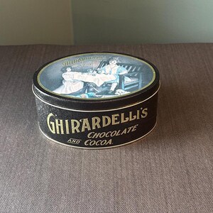 May include: A vintage Ghirardelli's Chocolate and Cocoa tin. The black tin features gold lettering and an oval lid with a painted scene of people. The tin is round and sits on a brown surface.