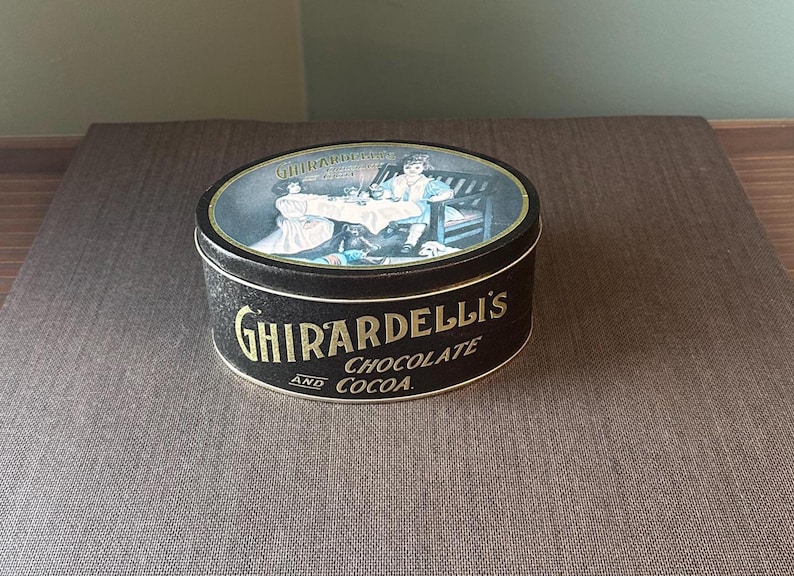 May include: A vintage Ghirardelli's Chocolate and Cocoa tin. The black tin features gold lettering and an oval lid with a painted scene of people. The tin is round and sits on a brown surface.