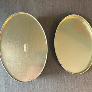 May include: Two oval-shaped, metallic containers with a silver rim. The containers have a gold-colored interior and are set against a gray background. The larger container has a small, light-colored mark on its surface.