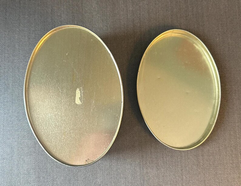 May include: Two oval-shaped, metallic containers with a silver rim. The containers have a gold-colored interior and are set against a gray background. The larger container has a small, light-colored mark on its surface.