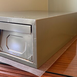 May include: An olive-green metal file cabinet with two drawers. The drawers have a rectangular design with a recessed panel and a pull-out handle. The word "Oxford" is embossed on the front. The cabinet is placed on a brown surface.