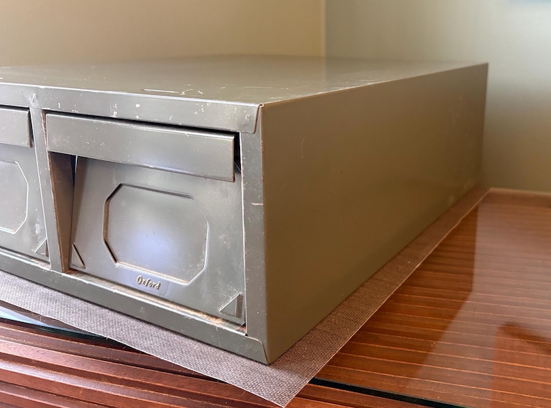 May include: An olive-green metal file cabinet with two drawers. The drawers have a rectangular design with a recessed panel and a pull-out handle. The word "Oxford" is embossed on the front. The cabinet is placed on a brown surface.