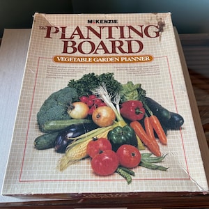 Puede incluir: La caja del planificador de jardín de vegetales Planting Board presenta una ilustración colorida de varios vegetales, incluyendo brócoli, berenjena, maíz, tomates y pimientos. La caja es blanca con texto rojo y negro.
