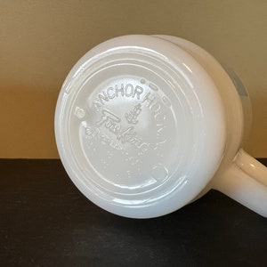 May include: White ceramic mug with a handle. The bottom of the mug has a circular logo that reads "Anchor Hocking Fire King Ovenware Made in USA".