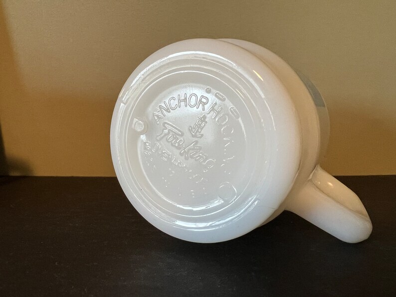 May include: White ceramic mug with a handle. The bottom of the mug has a circular logo that reads "Anchor Hocking Fire King Ovenware Made in USA".